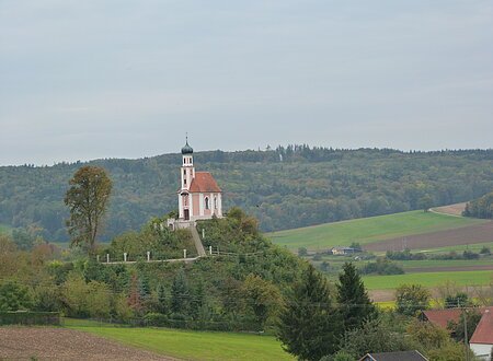 Auf einem Hügel mit steilen, bewaldeten Hängen steht eine kleine Kapelle mit rosa Fassade. Am Horizont ist ein bewaldeter Rücken zu erkennen.