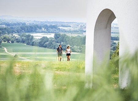 Zwei Personen wandern auf einem grünen Hügel, im Hintergrund eine weite Landschaft.