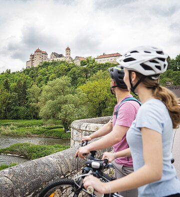 Radler auf der Steinernen Brücke in Harburg Zwei Personen mit Fahrrädern und Helmen stehen auf einer Brücke und blicken auf eine Burg auf einem Hügel.