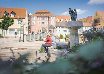 Personen sitzen auf einer Bank neben einem Brunnen auf einem Platz, im Hintergrund sind Gebäude mit roten Dächern.