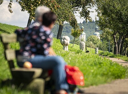 Verschwommene Personen auf einer Bank, im Hintergrund ein Weg mit Bäumen und Skulpturen.