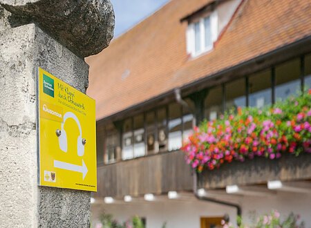 Lauschtour in Donauwörth Das gelbe Schild der Donauwörther Lauschtour. Im Hintergrund sieht man eine Familie und die alte Kaserne, ein altes Gebäude.