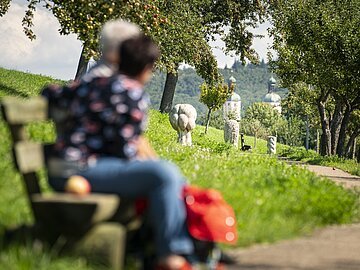 Bayerischer Jakobsweg in Wemding Verschwommene Personen auf einer Bank, im Hintergrund ein Weg mit Bäumen und Skulpturen.