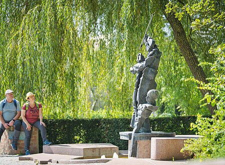 Zaubergeigenbrunnen in Donauwörth Zwei Personen sitzen auf einer Mauer neben einer abstrakten Skulptur im Freien, umgeben von Bäumen.