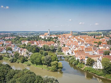 Luftaufnahme einer Stadt mit roten Dächern, Fluss und Brücke, umgeben von grüner Landschaft.