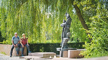 Zwei Personen sitzen auf einer Mauer neben einer abstrakten Skulptur im Freien, umgeben von Bäumen.