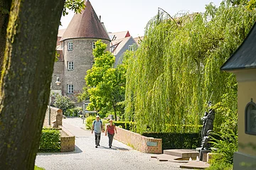 Zwei Personen spazieren auf einem gepflasterten Weg neben einer Backsteinmauer. Im Hintergrund ein Turm und Bäume.