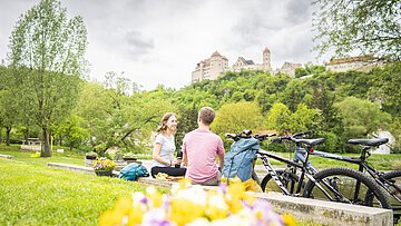 Ein Paar sitzt auf einer Wiese mit Fahrrädern, im Hintergrund die Harburg auf einem Hügel.