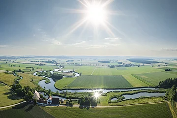 Landschaft mit einem sich schlängelndem Fluss, Feldern und Gebäuden, unter einem klaren Himmel mit strahlender Sonne.